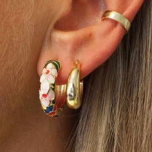 Pode incluir: Brincos de argola dourados com um design floral. O design floral apresenta esmalte branco, vermelho, azul e verde. Os brincos são usados na orelha.