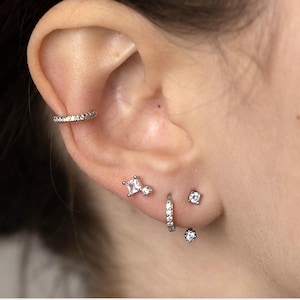 May include: A close-up of a person's ear with multiple silver earrings. The earrings are all set with small, clear cubic zirconia stones. The earrings include a hoop earring, a stud earring, and a small, square-shaped earring.