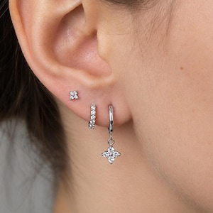 May include: A set of three silver-toned earrings consisting of a small four-petaled flower stud, a small hoop earring encrusted with clear stones, and a larger hoop earring featuring a dangling four-petaled flower charm with clear stones.