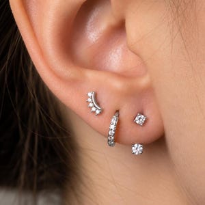 Pode incluir: Um conjunto de quatro brincos prateados usados numa única orelha. A coleção inclui um pino curvo com cinco pequenas pedras transparentes, uma pequena argola articulada com pedras transparentes e dois brincos individuais com pedras.