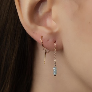 May include: A close-up of a gold-colored earring set. One earring features a chain with a vertical bar, while the other has a chain with three small, light blue beads. The earrings are worn in a person's ear.