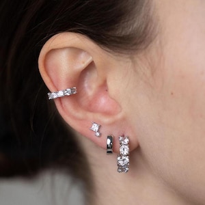 May include: A close-up of a person's ear with three silver earrings. The earrings are all adorned with small, clear cubic zirconia stones. The earrings are a hoop, a stud, and a cartilage cuff.