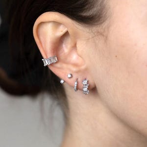 Könnte beinhalten: Nahaufnahme eines Ohrs mit mehreren silbernen Ohrringen. Die Ohrringe sind mit klaren Zirkonia-Steinen besetzt. Die Ohrringe haben die Form eines Kreises und einer Stange.