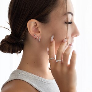 Pode incluir: Uma mulher vestindo um top cinza sem mangas com o cabelo puxado para tr&aacute;s em um coque. Ela est&aacute; usando v&aacute;rios pequenos brincos de argola de prata na orelha esquerda e um anel de prata com uma pequena pedra transparente no dedo anelar.
