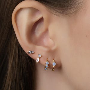 May include: A close-up of an ear adorned with multiple gold earrings. The earrings include a small stud with a clear stone, a teardrop-shaped earring with a clear stone, and two small hoop earrings with blue stones.