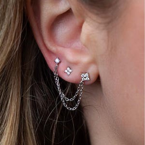 May include: A pair of silver earrings with small diamond-like stones. The earrings are connected by two silver chains.
