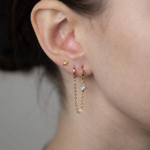 Puede incluir: Pendientes de estrella de oro con una cadena colgante y un dije de estrella. La cadena está conectada a un aro.