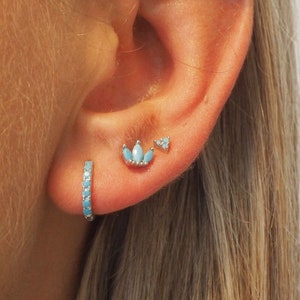 May include: A close-up of a person's ear with three earrings. The earrings are made of silver and have turquoise stones. The earrings are a small hoop, a small triangle, and a larger flower shape.