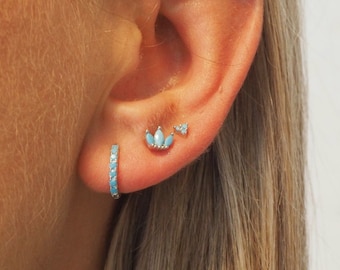 May include: A close-up of a person's ear with three earrings. The earrings are made of silver and have turquoise stones. The earrings are a small hoop, a small triangle, and a larger flower shape.