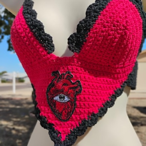 May include: A handmade, heart-shaped crochet top in bright pink with a black scalloped trim. The top features a patch of a red anatomical heart with an eye. The halter top is displayed on a mannequin.