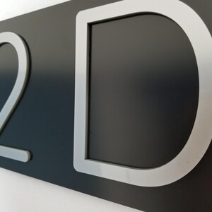 May include: A rectangular dark gray sign featuring the number 2 and the letter D in a large, silver-colored sans-serif font. The characters are raised and have a smooth, matte finish, contrasting against the dark background of the sign.
