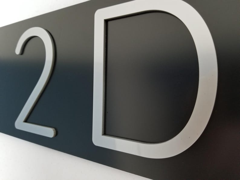 May include: A rectangular dark gray sign featuring the number 2 and the letter D in a large, silver-colored sans-serif font. The characters are raised and have a smooth, matte finish, contrasting against the dark background of the sign.