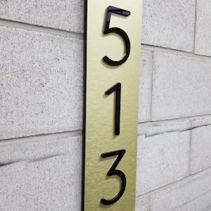 May include: A vertical rectangular house number sign featuring a brushed gold-tone metal plate with a black backing. The sign displays the raised black acrylic numbers 5, 1, and 3 arranged in a vertical column, mounted on a light gray textured wall.