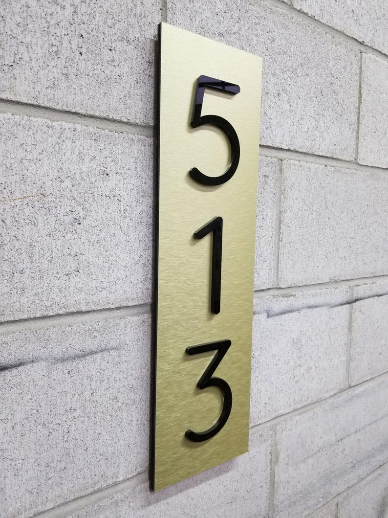 May include: A vertical rectangular house number sign featuring a brushed gold-tone metal plate with a black backing. The sign displays the raised black acrylic numbers 5, 1, and 3 arranged in a vertical column, mounted on a light gray textured wall.