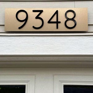 May include: A rectangular house number plaque featuring a brushed gold metallic finish with the black numbers 9348 centered on the surface. The plaque is mounted horizontally above a white door frame with a textured horizontal siding background.
