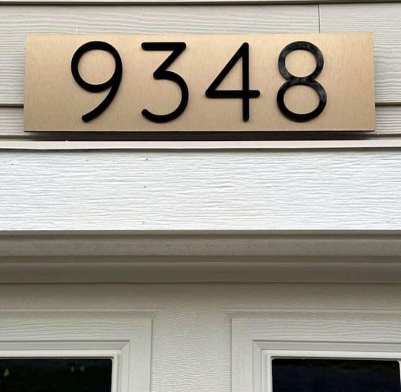 May include: A rectangular house number plaque featuring a brushed gold metallic finish with the black numbers 9348 centered on the surface. The plaque is mounted horizontally above a white door frame with a textured horizontal siding background.