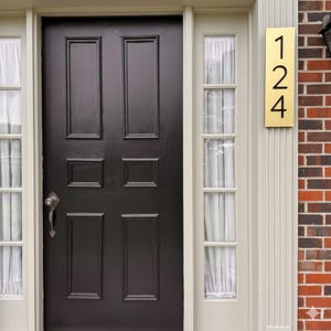 May include: A rectangular brass house number plaque featuring the black digits 1, 2, and 4 vertically aligned. The plaque is mounted on a cream-colored door frame next to a dark brown six-panel door with a silver handle and side windows.