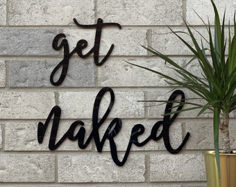 May include: Black cursive script wall decor with the words "get naked" on a white brick wall.
