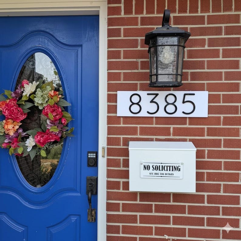 May include: A bright blue front door with an oval glass insert featuring a colorful floral wreath, a white rectangular house number sign displaying 8385, a black outdoor wall lantern, and a white mailbox with a No Soliciting sign attached.