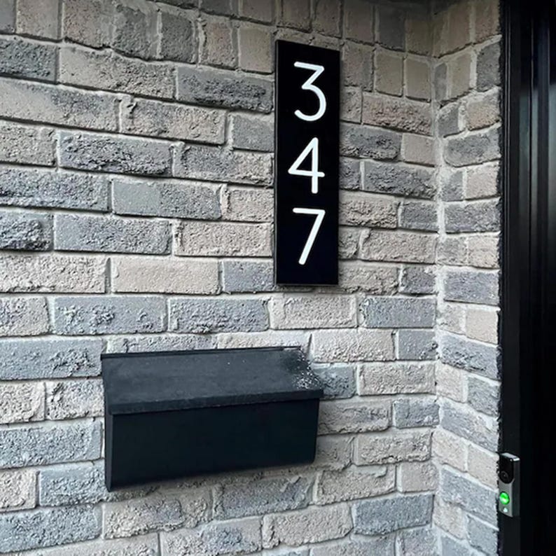 May include: A rectangular black house number sign featuring the white vertical digits 3, 4, and 7, mounted on a gray brick wall above a black metal mailbox. The sign displays the address number 347 in a clean, modern sans-serif font style.