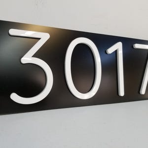 May include: A rectangular black house number sign featuring the white raised numerals 3017. The numbers are centered on the horizontal plaque and have a smooth, glossy finish that contrasts with the matte black background of the sign.