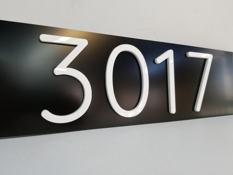 May include: A rectangular black house number sign featuring the white raised numerals 3017. The numbers are centered on the horizontal plaque and have a smooth, glossy finish that contrasts with the matte black background of the sign.