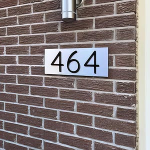 May include: A rectangular house number sign featuring the black digits 464 mounted on a smooth, brushed silver-toned metal plate. The sign is attached to a textured brown brick wall, positioned directly below a cylindrical silver light fixture.