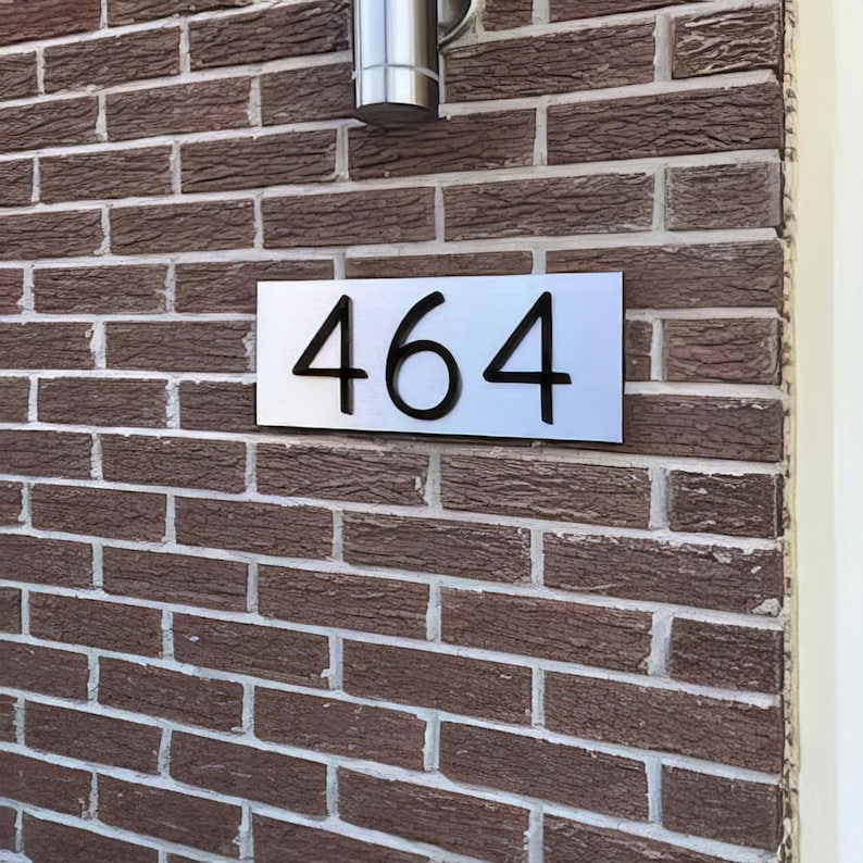 May include: A rectangular house number sign featuring the black digits 464 mounted on a smooth, brushed silver-toned metal plate. The sign is attached to a textured brown brick wall, positioned directly below a cylindrical silver light fixture.