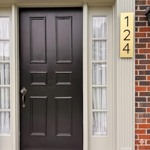 May include: A rectangular brass house number plaque featuring the black digits 1, 2, and 4 vertically aligned. The plaque is mounted on a cream-colored door frame next to a dark brown six-panel door with a silver handle and side windows.
