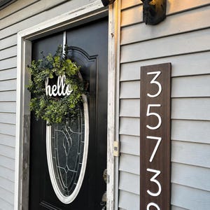 May include: A vertical rectangular dark wood address sign with white numbers 35738 mounted on light gray siding next to a black door. A green eucalyptus wreath with a white script hello sign hangs on the oval glass window of the door.