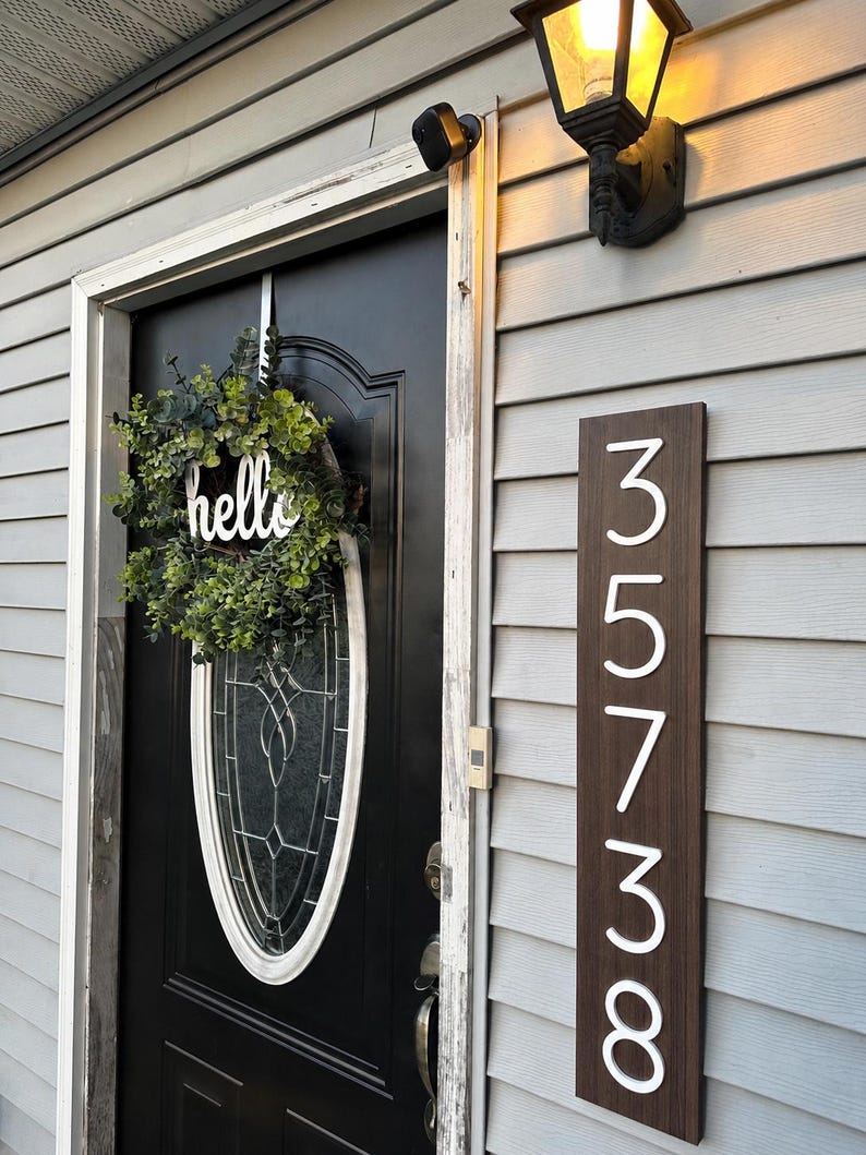May include: A vertical rectangular dark wood address sign with white numbers 35738 mounted on light gray siding next to a black door. A green eucalyptus wreath with a white script hello sign hangs on the oval glass window of the door.