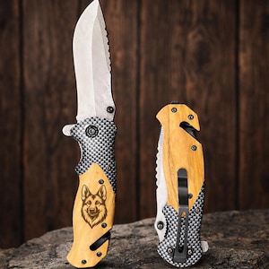 May include: Two folding knives with silver blades and wooden handles. One knife features a German Shepherd engraving. The handles have a black and silver checkered pattern. The knives are displayed on a stone surface against a wooden background.