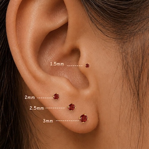 May include: Close-up of an ear with four red gemstone stud earrings in varying sizes: 1.5mm, 2mm, 2.5mm, and 3mm. The earrings are set in gold. The ear is shown against a blurred background.