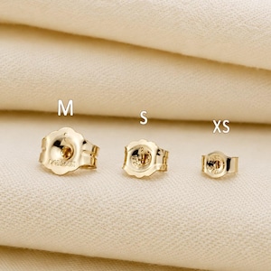 May include: Three gold-colored earring backs in varying sizes, labeled M, S, and XS from left to right. Each piece features a scalloped edge design and a central opening for an earring post, showcasing a metallic finish and a circular structure.