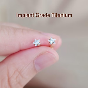 May include: A pair of gold-coloured star-shaped earrings, each with a light blue gemstone centre. The earrings are held in a person's hand. The text "Implant Grade Titanium" is displayed above the earrings.