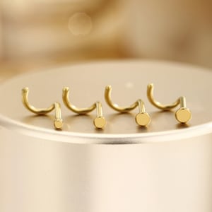 Peut inclure: Plusieurs clous de nez dorés sont présentés, chacun avec une tige incurvée et une petite extrémité circulaire. Les clous sont disposés sur une surface dorée. L'image a une esthétique minimaliste, mettant l'accent sur le design et la couleur des bijoux.