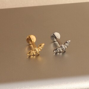 May include: Two bat-shaped earrings, one gold-toned and one silver-toned, are displayed. Each earring features a small, flat disc and a post for piercing. The bat wings are detailed, and the earrings are set against a neutral background.