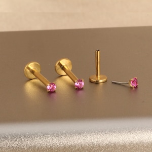 Może przedstawiać: Złote kolczyki do piercingu warg z r&oacute;żowymi kamieniami szlachetnymi. Kolczyki mają płaską, dyskową podstawę i prosty słupek. Jeden kolczyk jest odłączony, pokazując słupek i kamień szlachetny. Biżuteria jest prezentowana na neutralnej powierzchni.