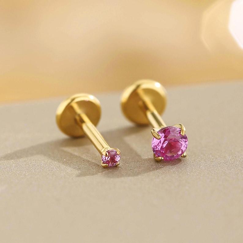 K&ouml;nnte beinhalten: Zwei goldfarbene Ohrstecker mit rosa Edelsteinen. Ein Ohrring hat einen kleinen rosa Edelstein, der andere einen gr&ouml;&szlig;eren, runden rosa Edelstein. Die Ohrringe sind vor einem neutralen Hintergrund platziert.