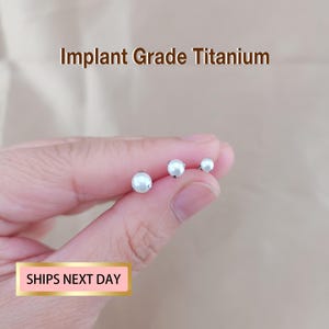 May include: Three white pearl stud earrings in varying sizes are displayed. The text "Implant Grade Titanium" is above the earrings. The earrings are held in a hand. The text "SHIPS NEXT DAY" is in a pink box.