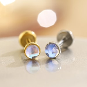 Könnte beinhalten: Zwei Ohrstecker mit irisierenden, runden Edelsteinen. Ein Ohrring hat eine goldene Fassung, der andere eine silberne Fassung. Die Edelsteine reflektieren eine Reihe von Farben, darunter Blau, Rosa und Gelb. Die Ohrringe sind auf einer reflektierenden Oberfläche.