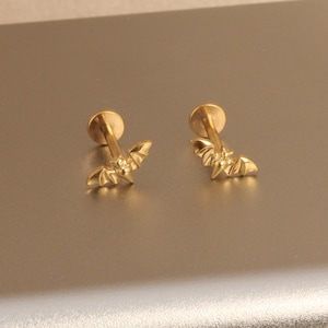 May include: A pair of gold-colored bat-shaped stud earrings. Each earring features a small, detailed bat design with outstretched wings, attached to a round post. The earrings are displayed on a neutral, light-colored surface.