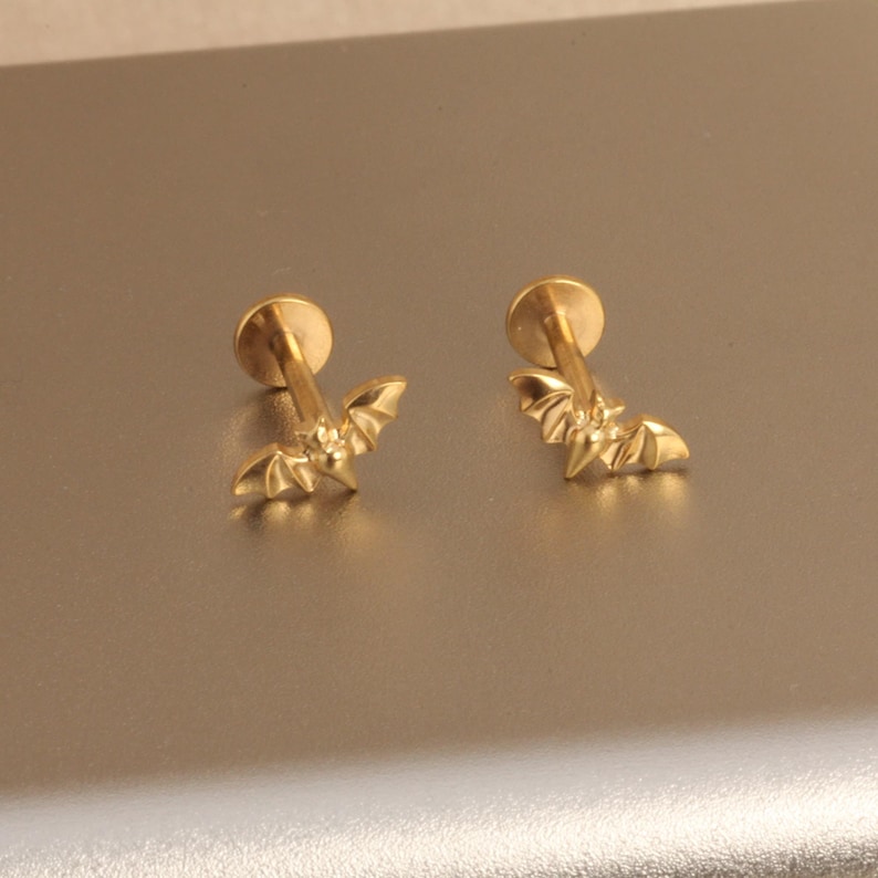 May include: A pair of gold-colored bat-shaped stud earrings. Each earring features a small, detailed bat design with outstretched wings, attached to a round post. The earrings are displayed on a neutral, light-colored surface.