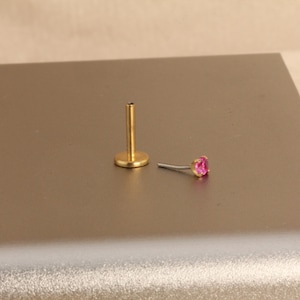 Może przedstawiać: Dwa elementy biżuterii. Jednym z nich jest złoty, płaski kolczyk z trzpieniem. Drugi to mały, srebrny kolczyk z r&oacute;żowym kamieniem szlachetnym. Biżuteria znajduje się na neutralnej powierzchni.