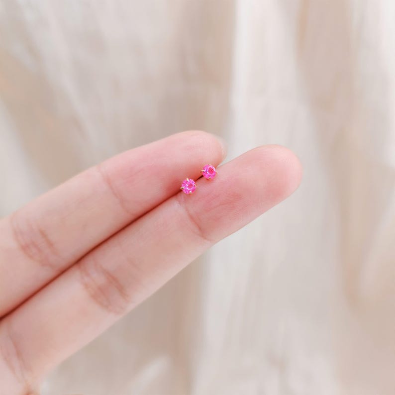 K&ouml;nnte beinhalten: Ein Paar kleiner, leuchtend rosa Ohrstecker. Jeder Ohrring hat einen runden, facettierten Edelstein. Die Ohrringe werden auf einem Finger vor einem weichen, neutralen Hintergrund pr&auml;sentiert.