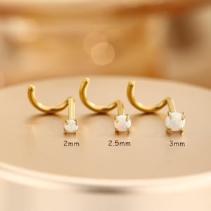 May include: Three gold-tone nose studs with white opal-like stones in prong settings. The studs are arranged in a row with labels indicating sizes of 2mm, 2.5mm, and 3mm. Each piece features a curved post design for secure placement.
