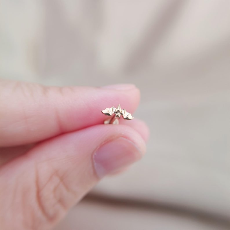 May include: A small, gold-colored bat-shaped earring stud. The earring features a detailed design of a bat with outstretched wings. The earring is held between fingers against a blurred background.