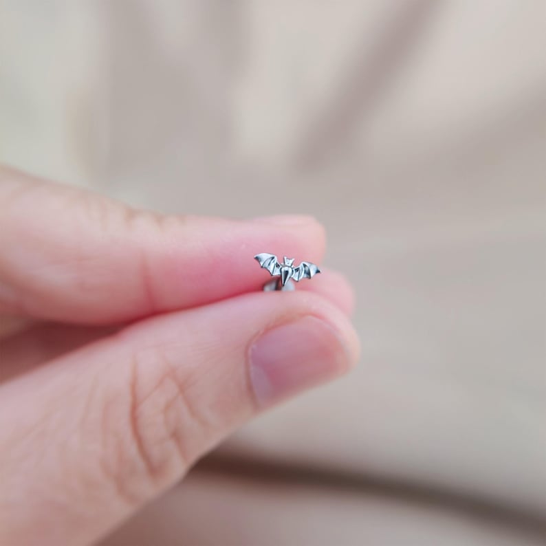 May include: A small, silver-colored bat-shaped earring. The earring features a detailed design of a bat with outstretched wings. The earring is held between fingers against a blurred background.