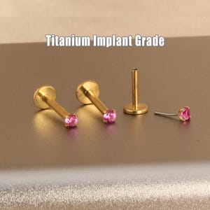 May include: Gold-colored lip piercing jewelry with a pink gemstone. The image shows several pieces, including a post and two complete pieces. The text "Titanium Implant Grade" is displayed above the jewelry.