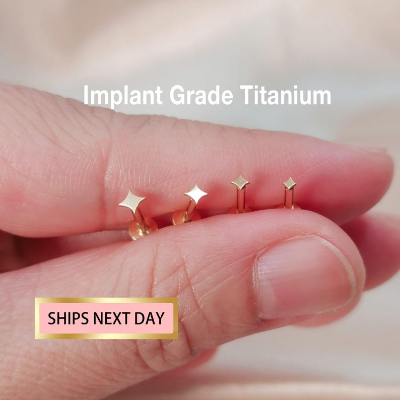 May include: Three gold titanium stud earrings with a diamond shape. The earrings are shown on a hand, with the text "Implant Grade Titanium" above the earrings.